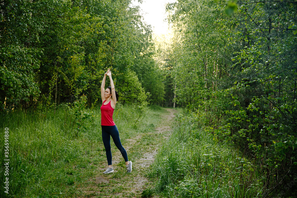 Young woman warming up at forest road before jogging. Attractive girl stretching for running exercises. Beautiful curly sporty girl doing fitness outdoor at summer day. Healthy lifestyle concept Young woman warming up at forest road before jogging. Attractive girl stretching for running exercises. Beautiful curly sporty girl doing fitness outdoor at summer day. Healthy lifestyle concept