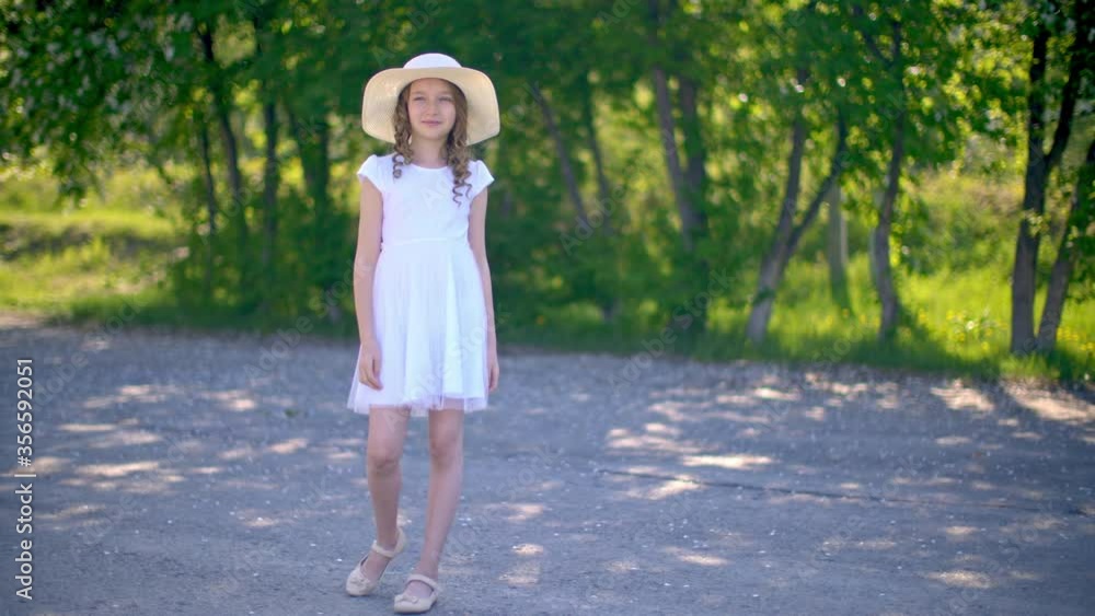 Young girl in white dress and hat standing on park alley on green trees background. Beautiful girl in summer park at sunny day. Romantic model in spring garden Young girl in white dress and hat standing on park alley on green trees background. Beautiful girl in summer park at sunny day. Romantic model in spring garden