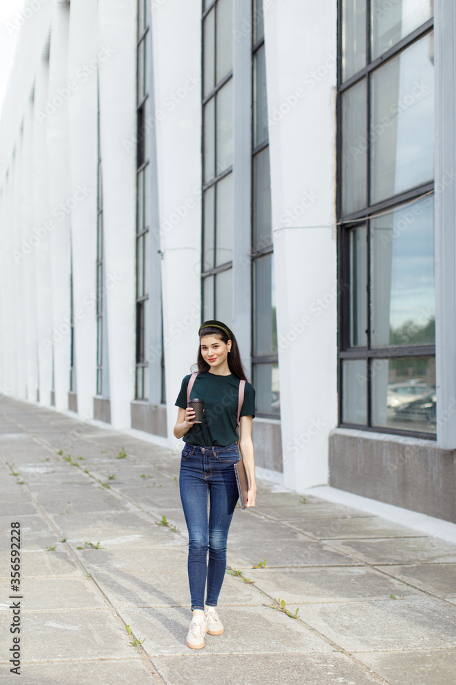Beautiful young brunette woman carrying backpack and drinking takeaway coffee while walking outdoors