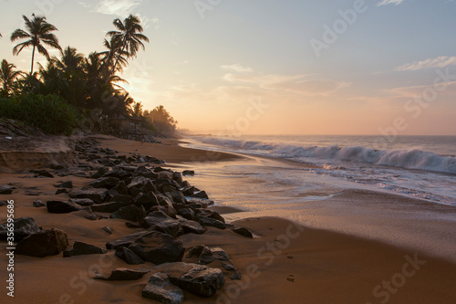 Beautiful postcard landscape shot of the Pitiwella beach (Sri Lanka) on sunrise with orange sand, green palm trees, brown rocks, the Indian ocean with big white waves and the sun on the horizon photo