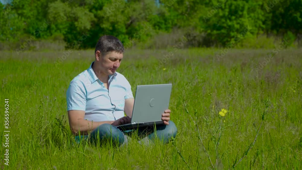 Man Works behind a Laptop Sitting on the Grass At The Sunny Day. Freelancer Remote Work Concept. 4K Man Works behind a Laptop Sitting on the Grass At The Sunny Day. Freelancer Remote Work Concept. 4K
