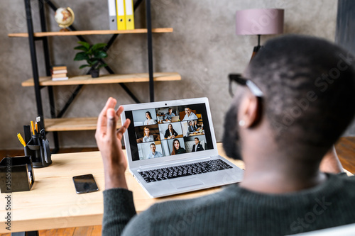 Confident African-American male worker talking online with coworkers, back view of black guy speaks and gestures to many people on video screen. Remote work, virtual meeting photo