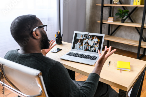 Virtual meeting with many people together. African-American young guy talking online with employees via video connection. Multiracial team. Back view photo