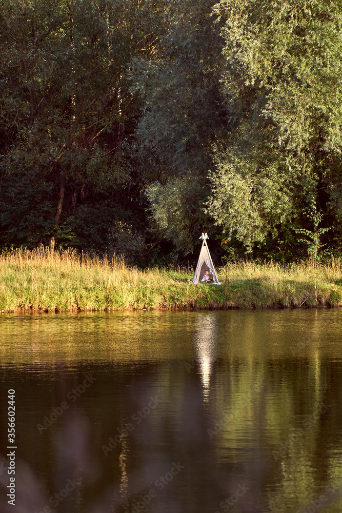 Fototapeta premium Pond on a sunny day. Wigwam on the other shore. Lake in the forest. Green fairy tale. High quality photo