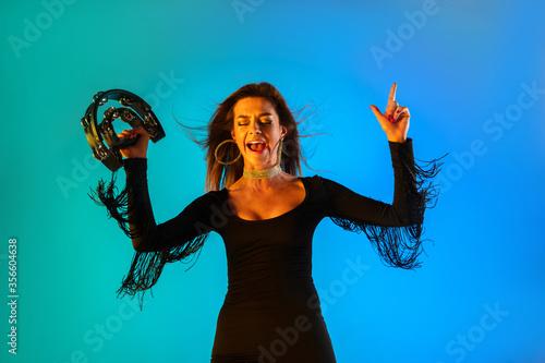 Caucasian female singer with tambourin isolated on blue studio background in neon light. Beautiful female model in black dress. Concept of human emotions, facial expression, ad, music, art, festival. photo