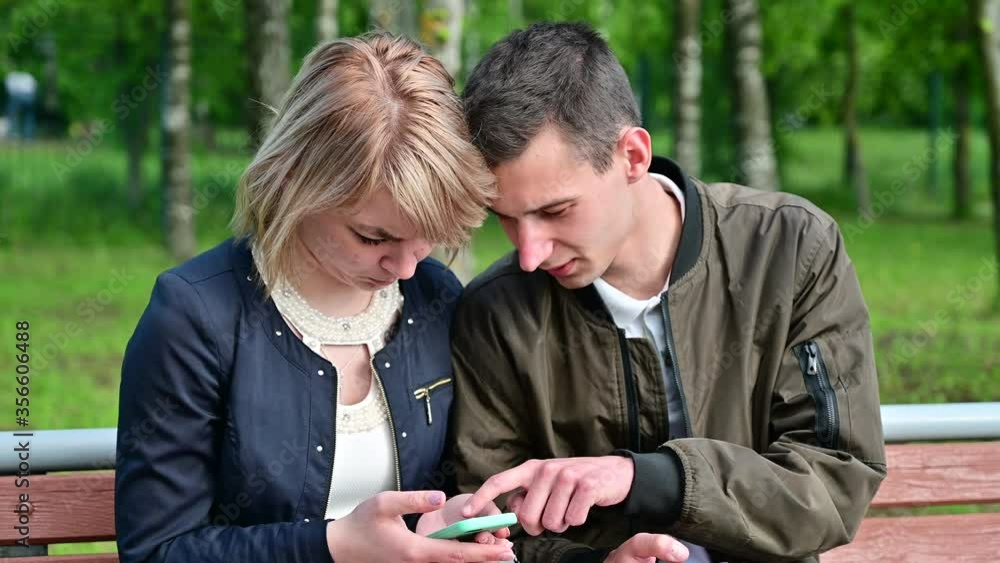 Young couple use a smartphone while sitting on a bench in the park. The concept of family communication. Relationship problems. Young couple use a smartphone while sitting on a bench in the park. The concept of family communication. Relationship problems.