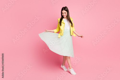Full length photo of funky lady walking down street good mood raise green long wavy skirt wear trend yellow leather jacket sneakers isolated pastel pink color background