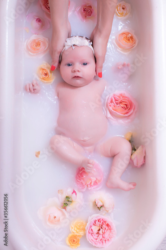 Happy kid girl takes a milk bath with petals. Bouquets of pastel rose flowers. Infant bathing. Hygiene and care for young children.
