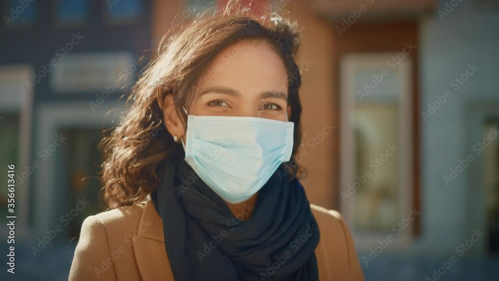 Portrait of a Beautiful Dark Haired Young Woman Wearing Protective Medical Face Mask and Standing on the Street. Safe and Happy Woman Practicing Social Distancing, Quarantine. Blurred City Background Portrait of a Beautiful Dark Haired Young Woman Wearing Protective Medical Face Mask and Standing on the Street. Safe and Happy Woman Practicing Social Distancing, Quarantine. Blurred City Background