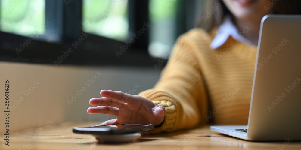 Cropped image of an office woman is charging a smartphone with a wireless charger while typing on a computer laptop. - obrazy, fototapety, plakaty 