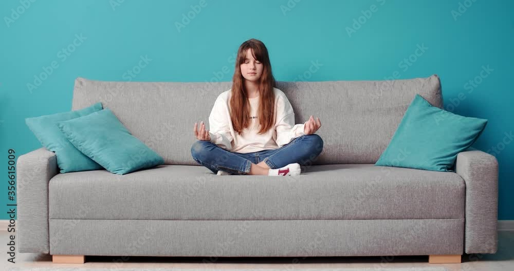 Pretty young girl meditating on grey couch with closed eyes. Teenager girl with dark hair relaxing at home during free time. Pretty young girl meditating on grey couch with closed eyes. Teenager girl with dark hair relaxing at home during free time.