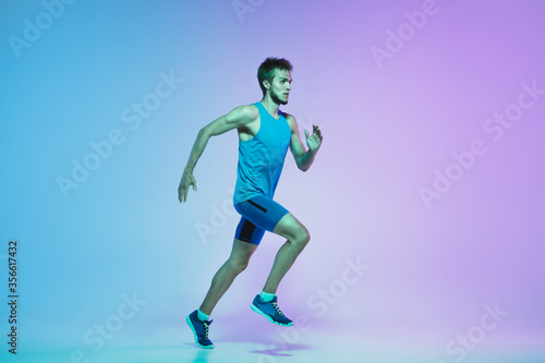 Portrait of active young caucasian man running, jogging on gradient studio background in neon light. Professional sportsman training in action and motion. Sport, wellness, activity, vitality concept.