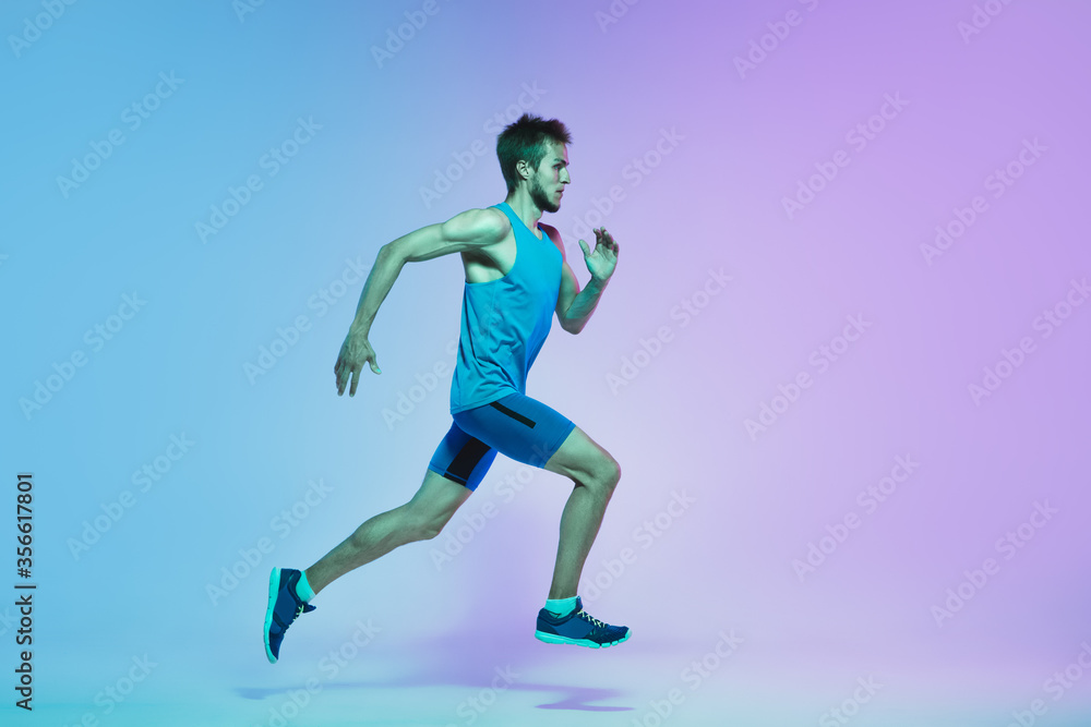 Portrait of active young caucasian man running, jogging on gradient studio background in neon light. Professional sportsman training in action and motion. Sport, wellness, activity, vitality concept.
