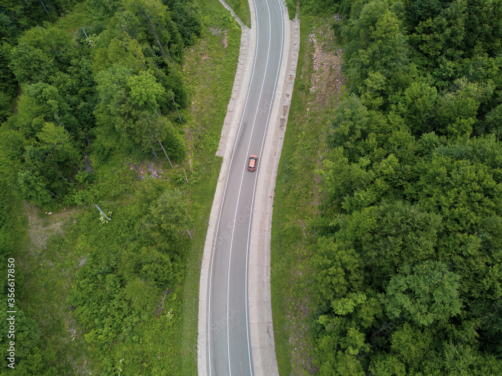 Drone point of view: aerial view flying over two lane countryside forest road with orange car moving green trees of dense woods growing both sides.