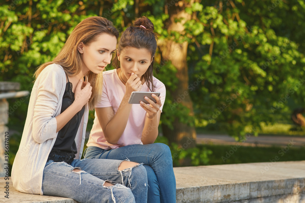 Girlfriends look in phone, view photos with vacation, recall the nice moments. Beautiful women in ordinary clothes sitting in the Park. Women are surprised by news, shocked by content and information Girlfriends look in phone, view photos with vacation, recall the nice moments. Beautiful women in ordinary clothes sitting in the Park. Women are surprised by news, shocked by content and information