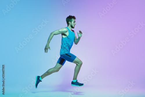 Portrait of active young caucasian man running  jogging on gradient studio background in neon light. Professional sportsman training in action and motion. Sport  wellness  activity  vitality concept.