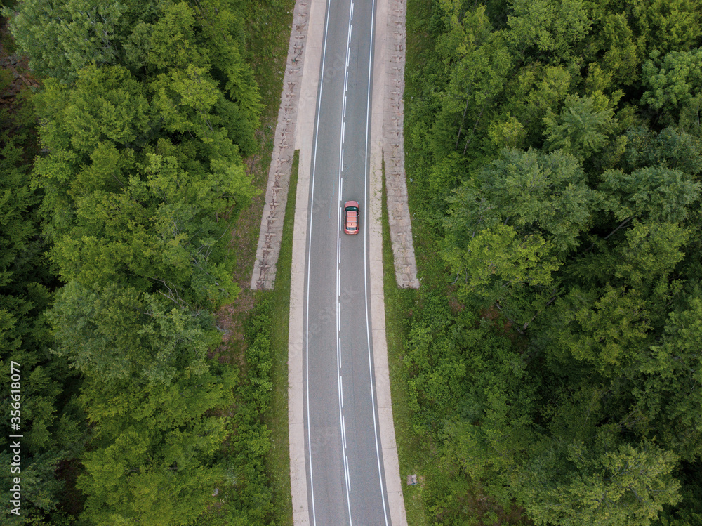 Drone point of view: aerial view flying over two lane countryside forest road with orange car moving green trees of dense woods growing both sides. Drone point of view: aerial view flying over two lane countryside forest road with orange car moving green trees of dense woods growing both sides.
