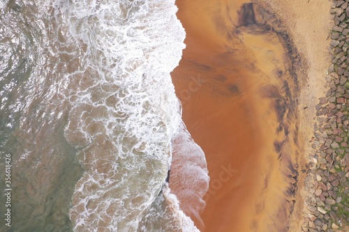 aerial drone bird view shot of the sea shore with yellow sand, black rocks, large white waves and foam crashing on the beach forming beautiful textures, patterns, shapes. Sri Lanka