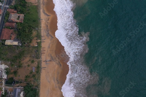 natural aerial drone bird view day shot of the sea shore with a road, beautiful villas, green trees, yellow sand, turquoise blue water, big waves and foam crashing on the beach. Pitiwella, Sri Lanka © Liza