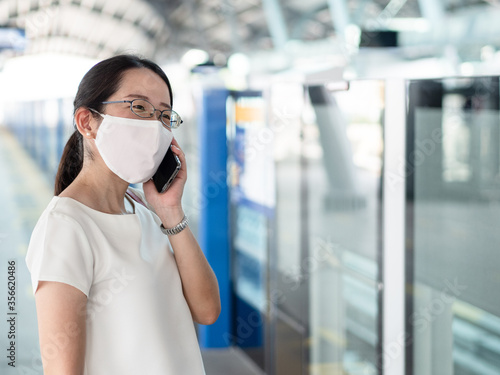 Beautiful Asian women wearing disposable medical face mask, using smartphone while waiting for metro at train station platform, as new normal trend and self-protection against Covid19 infection.