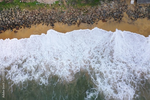 aerial drone bird view shot of the sea shore with yellow sand, black rocks, large white waves and foam crashing on the beach forming beautiful textures, patterns, shapes. Sri Lanka