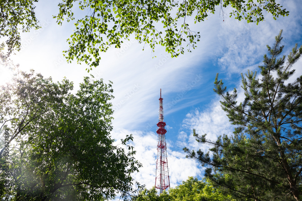 red tall television tower on a background of blue sky and clouds and green trees