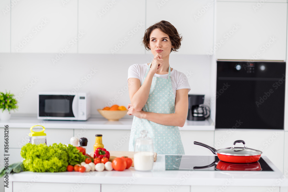 Photo of beautiful housewife holding arm on chin remind recipe minded enjoy weekend morning cooking tasty dinner family meeting wear apron t-shirt stand modern kitchen indoors Photo of beautiful housewife holding arm on chin remind recipe minded enjoy weekend morning cooking tasty dinner family meeting wear apron t-shirt stand modern kitchen indoors