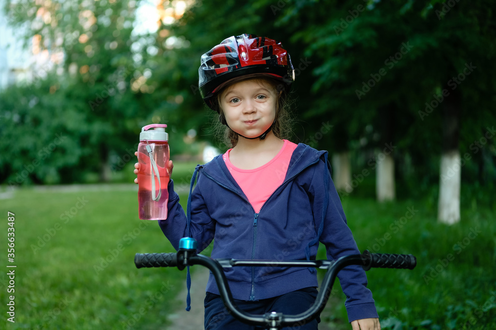 Obraz premium Little girl drinking water by the bike. Child in helmet riding a bicycling.