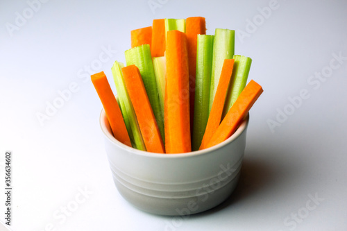 close up filled frame isolated shot of party snack food. A white bowl of crunchy orange carrot and juicy green celery sticks on a white background