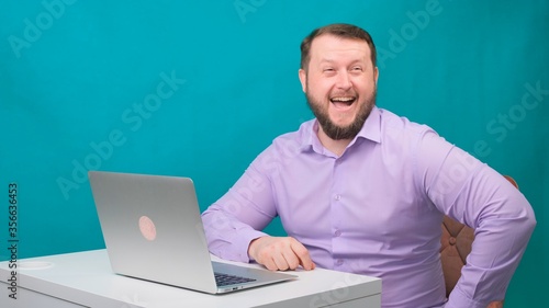 Young happy businessman smiling and speak into the camera. Portrait of a male laughing while looking at his laptop. Man working at his desk in the office