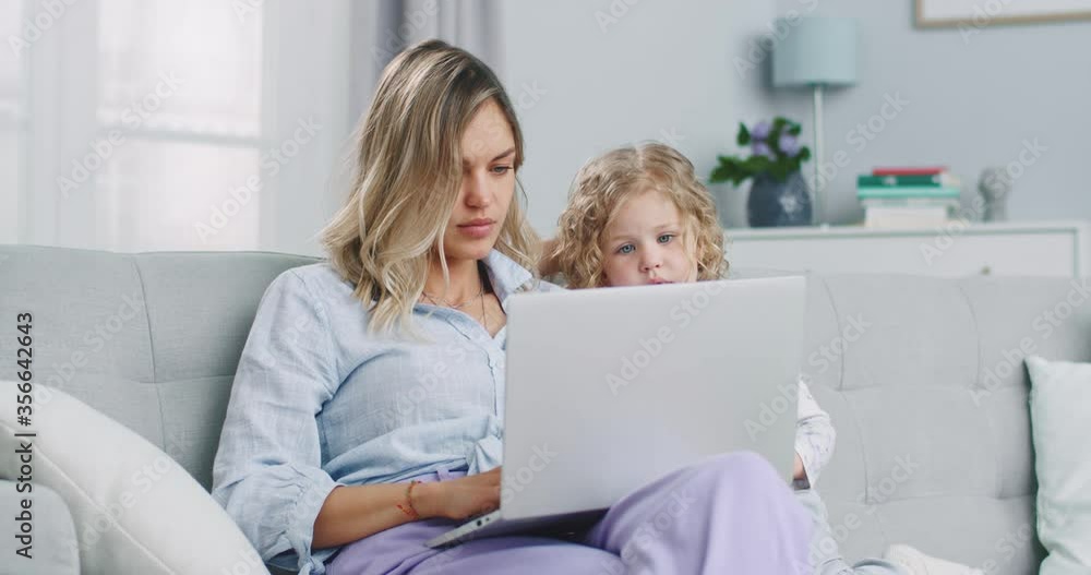 Middle plan of caucasian mother working from home, having a work video call, daughter playing nearby. Stay home, quarantine remote work. Concept of family, parenthood, togetherness. Middle plan of caucasian mother working from home, having a work video call, daughter playing nearby. Stay home, quarantine remote work. Concept of family, parenthood, togetherness.