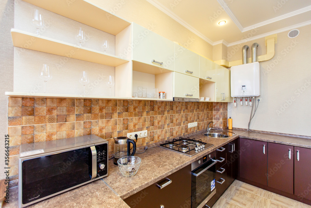 Kitchen in the apartment with a fresh renovation. Kitchen furniture in beige tones, brown tiles. Kitchen appliances: refrigerator, stove, microwave, gas boiler Kitchen in the apartment with a fresh renovation. Kitchen furniture in beige tones, brown tiles. Kitchen appliances: refrigerator, stove, microwave, gas boiler