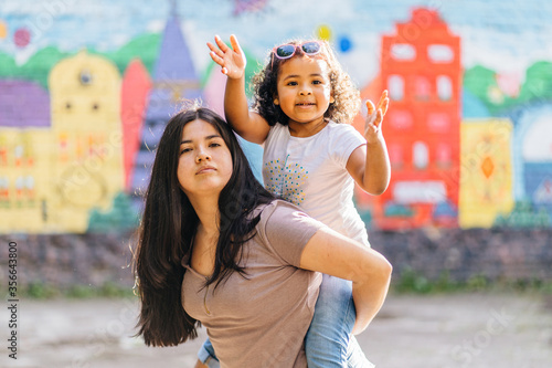 Happy family piggyback. Parenthood, childhood, love and happiness concept. Single mixed race mother playing with her cute curly preschooler daughter outdoor in city street, bright blurred background.