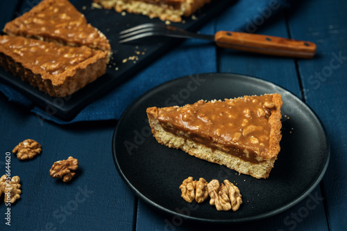 A piece of tart on the black plate and chopped walnut caramel pie on the wooden table © Grigoriy