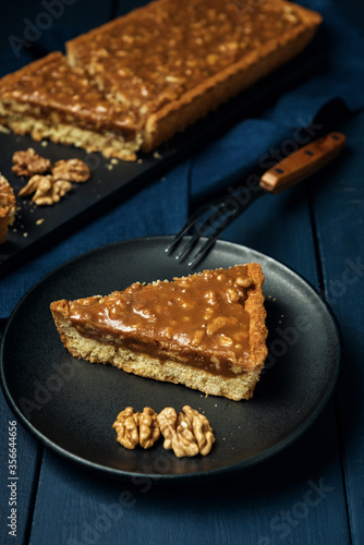 A piece of tart on the black plate and chopped walnut caramel pie on the wooden table © Grigoriy