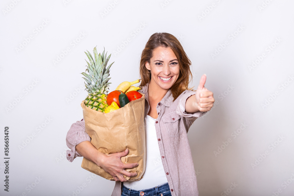 Carrying a healthy bag. Cropped image of beautiful young woman in apron holding paper shopping bag full of fresh vegetables and smiling while showing thumbs up Carrying a healthy bag. Cropped image of beautiful young woman in apron holding paper shopping bag full of fresh vegetables and smiling while showing thumbs up