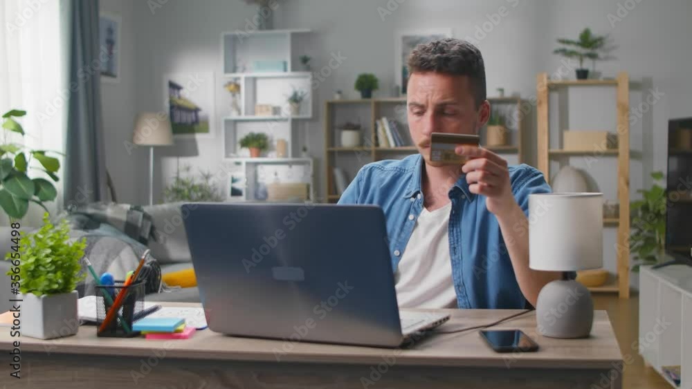 Young man makes an online purchase using a laptop and a bank card in his living room Young man makes an online purchase using a laptop and a bank card in his living room