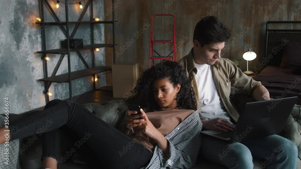 Couple moving to new apartment. He working at laptop, she using smartphone. Relaxing together on gray couch in room with cardboard boxes. Close up Couple moving to new apartment. He working at laptop, she using smartphone. Relaxing together on gray couch in room with cardboard boxes. Close up