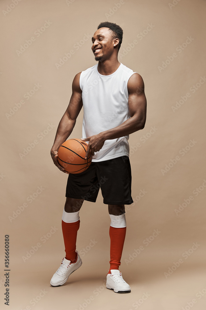 full length photo of a black athletic basketball player in the studio on a beige background wearing a white T-shirt, black shorts, red long socks and white sneakers, he holds the ball and smiles