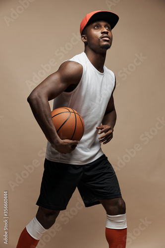 photo of a dark-skinned athletic basketball player in studio on a beige background posing with a ball, wearing a white t-shirt, red cap, black shorts and he is looking up © monchak