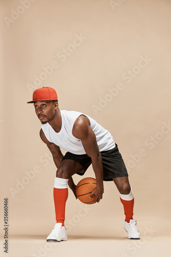 Fototapeta Naklejka Na Ścianę i Meble -  full length photo of a dark-skinned athletic basketball player in studio on a beige background posing with a ball, wearing a white t-shirt, black shorts, red long socks and a cap and white sneakers