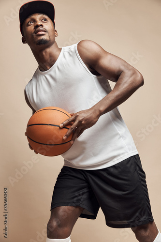 photo of a dark-skinned athletic basketball player in studio on a beige background posing with a ball, wearing a white t-shirt, black shorts, red cap and he is looking up © monchak