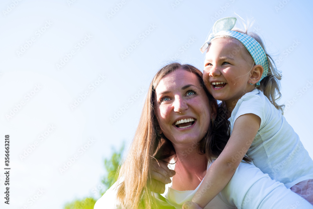 Cute little daughter on a piggy back ride with her mother. Happy mom and girl playing having fun outdoor in park at summer together on the green grass. Family lifestyle Cute little daughter on a piggy back ride with her mother. Happy mom and girl playing having fun outdoor in park at summer together on the green grass. Family lifestyle