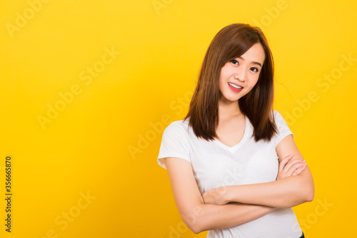 Asian happy portrait beautiful cute young woman teen standing wear t-shirt smile her confidence with crossed arms looking to camera isolated, studio shot on yellow background with banner copy space