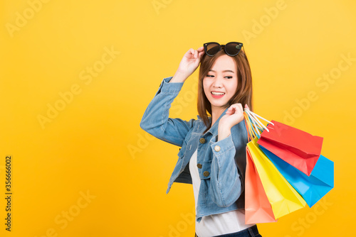 Asian happy portrait beautiful cute young woman teen smiling standing with sunglasses excited holding shopping bags multi color looking bags isolated, studio shot yellow background with copy space