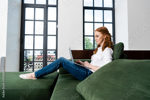 Side view of red haired freelancer using laptop on sofa