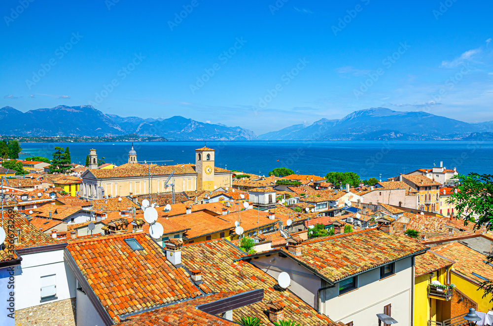 Aerial panoramic view of Desenzano del Garda town with bell tower of Duomo di Santa Maria Maddalena Cathedral church, red tiled roof buildings, Garda Lake, mountain range, Lombardy, Northern Italy