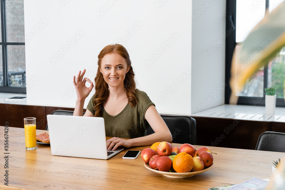 Selective focus of smiling woman showing ok gesture near laptop, smartphone and breakfast on table Selective focus of smiling woman showing ok gesture near laptop, smartphone and breakfast on table
