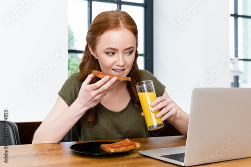 Woman eating toast and holding glass of orange juice near laptop on wooden table