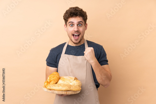Male baker holding a table with several breads isolated on beige background celebrating a victory